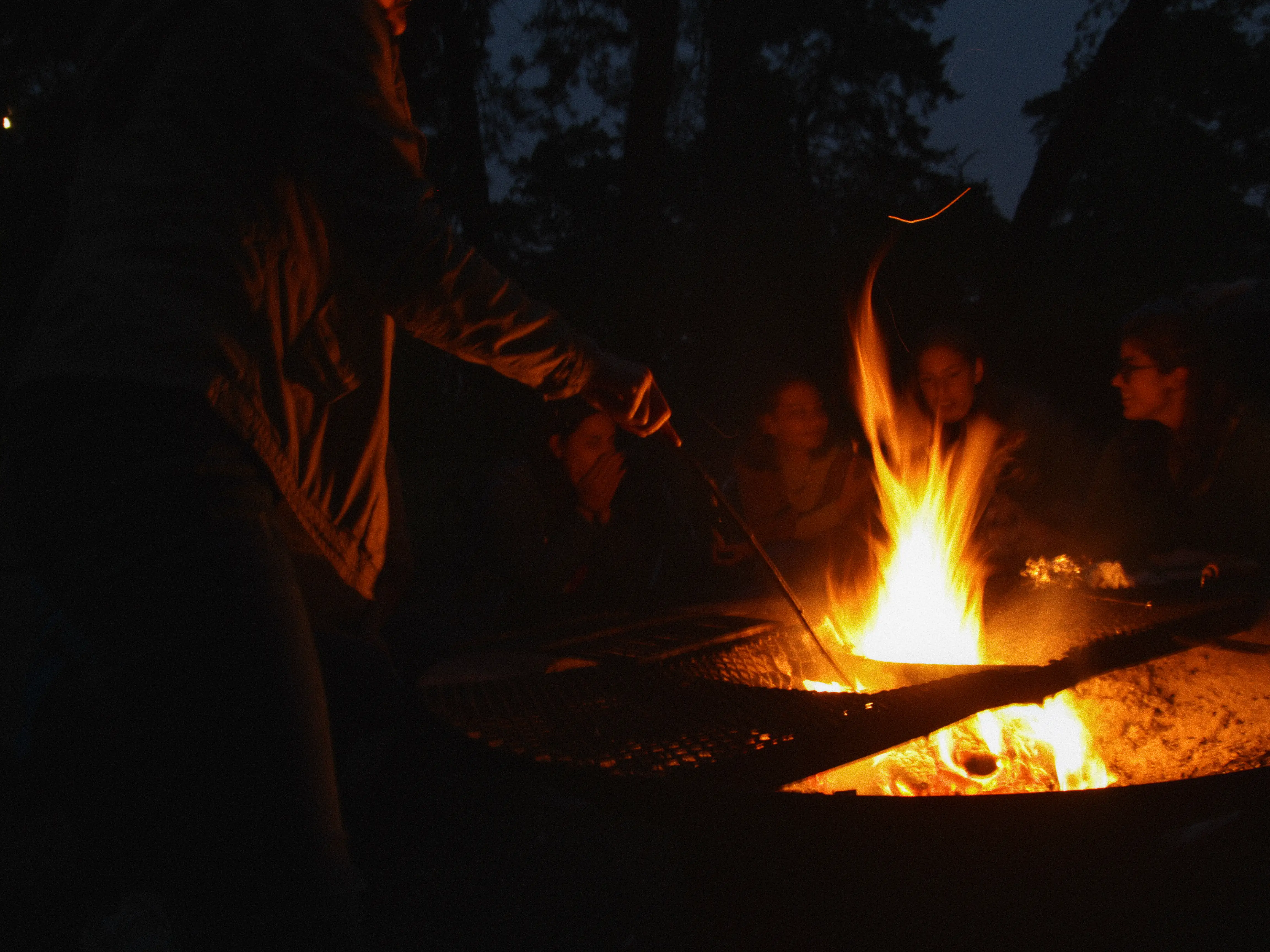 A person managing a campfire with people sitting around it, in a dark blue forested background.