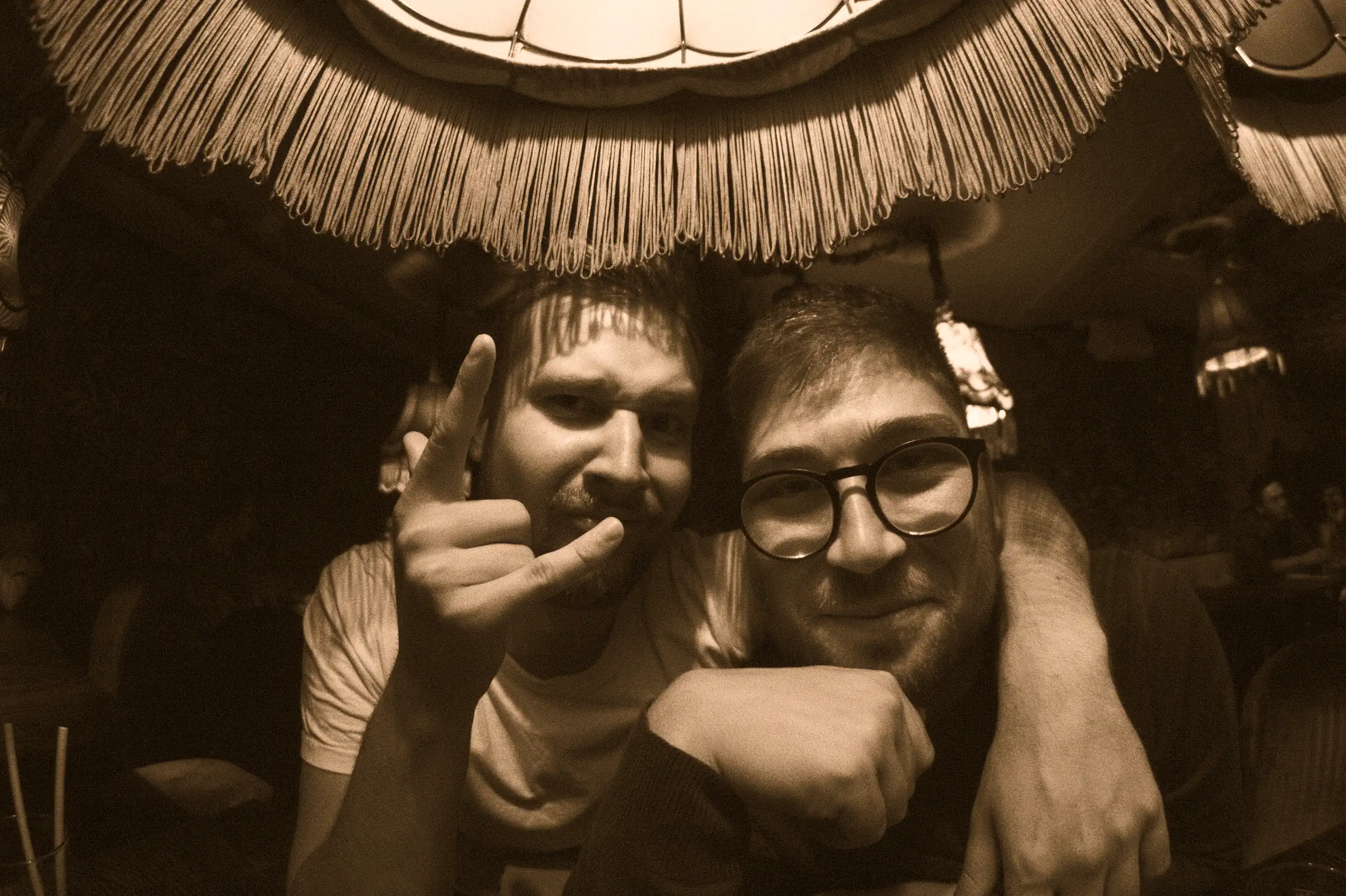 Two men making finger signs and smiling at the camera, sitting in a dimly lit room