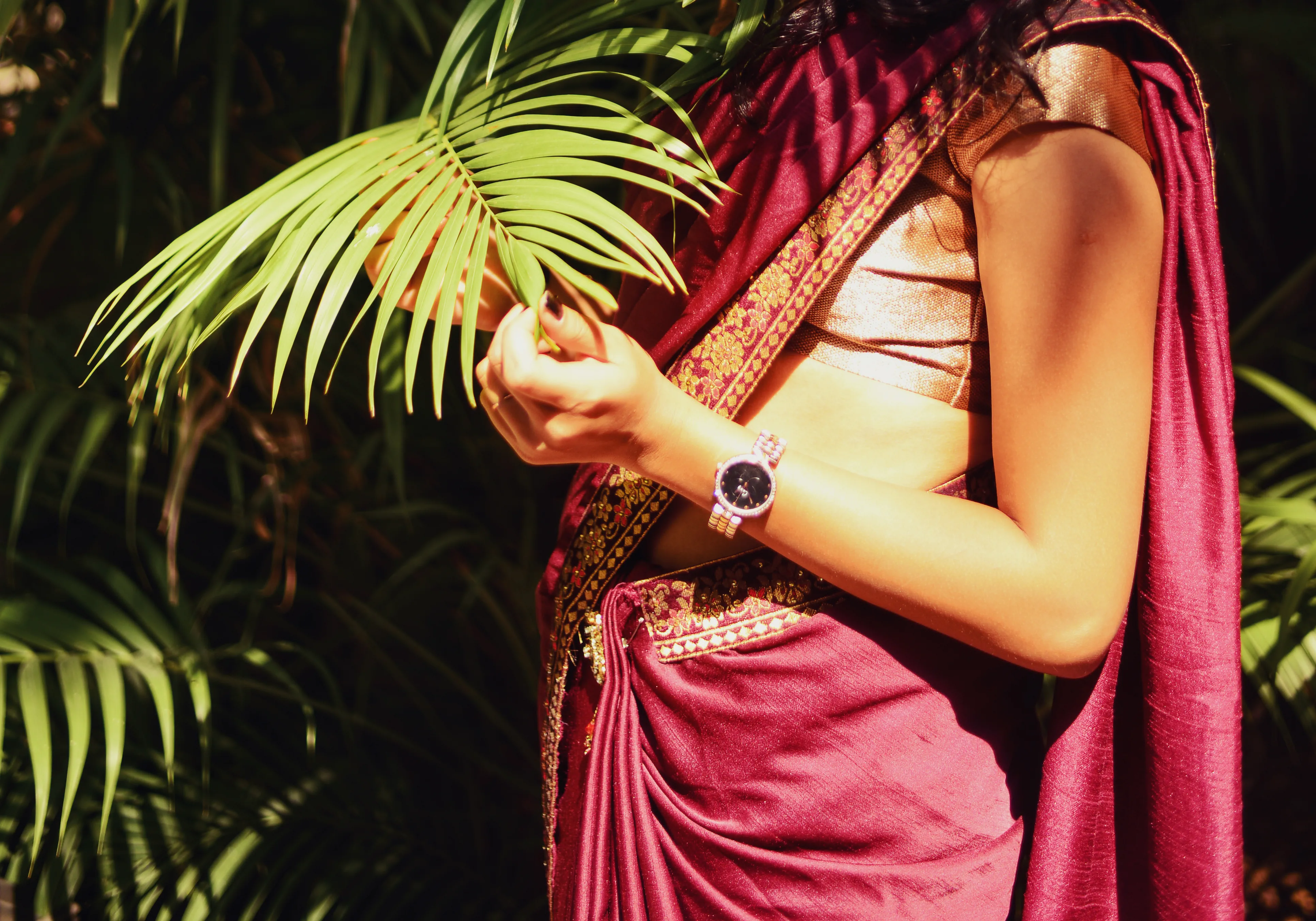 A woman dressed in a maroon Saree, fiddling with pam leaves with the sun falling on her body