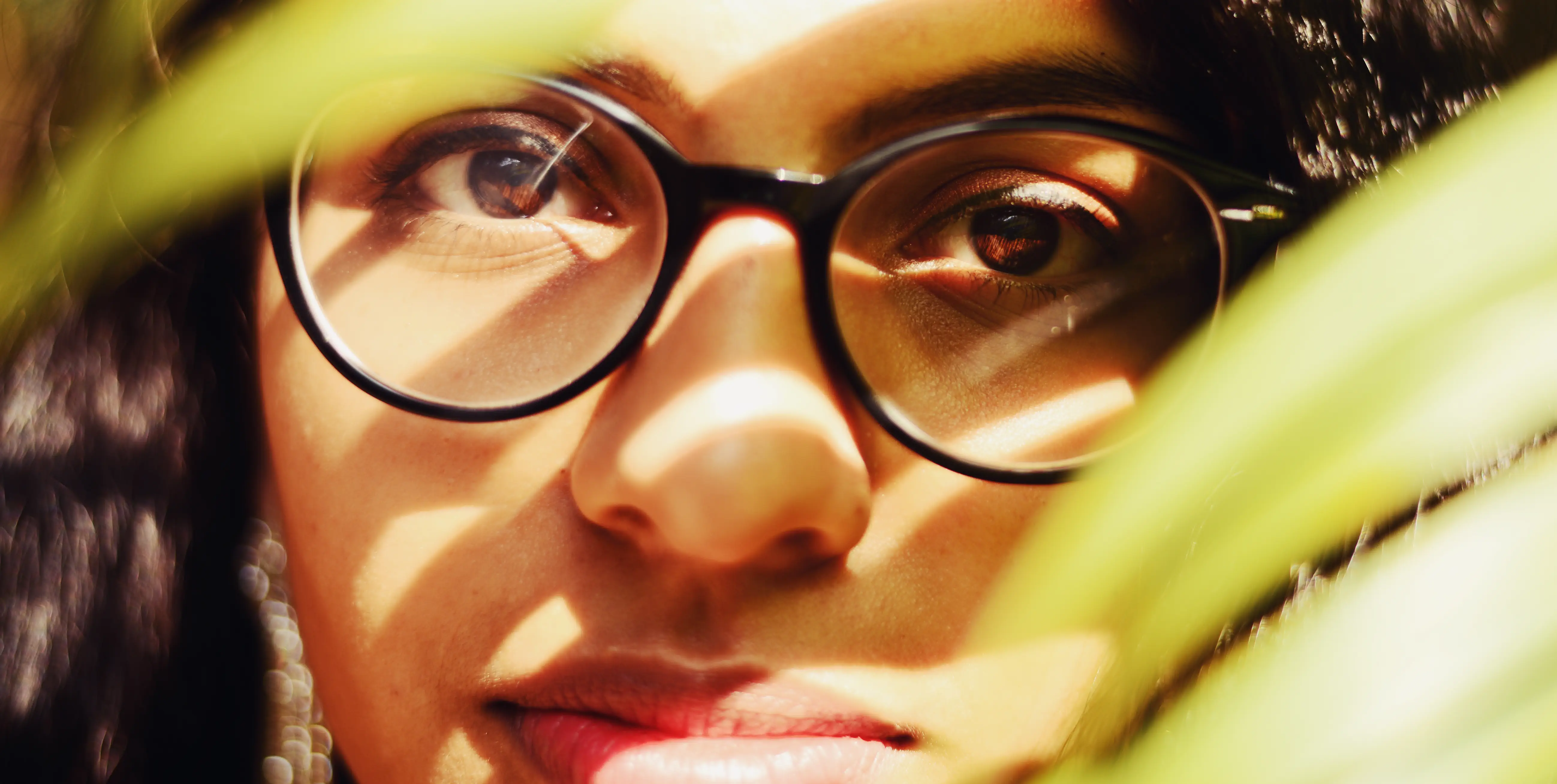 A woman looking at the camera through palm leaves with sunlight falling onto the iris of her eyes